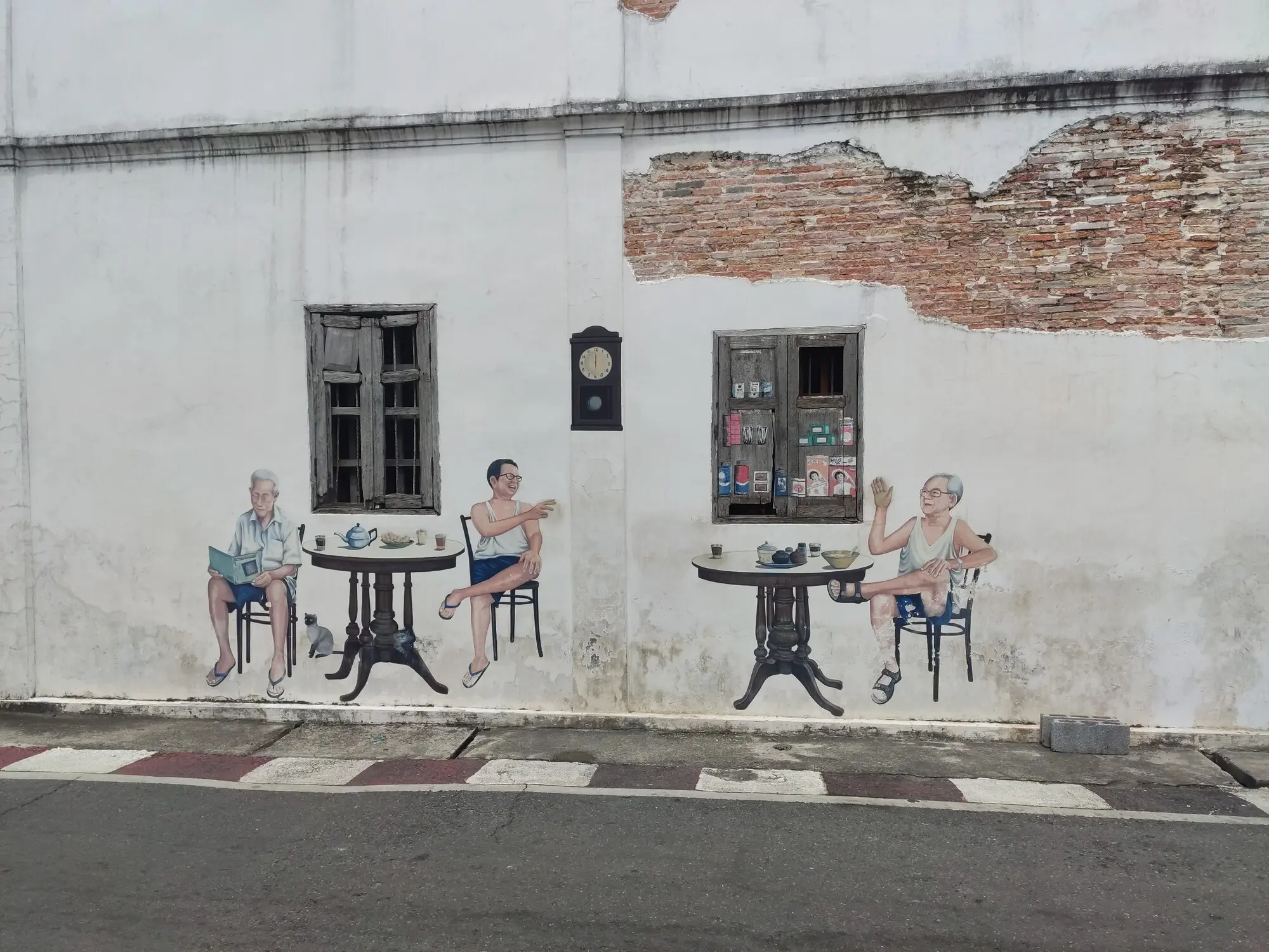 Фотография уличной стены со стрит-артом. Нарисованы три человека, сидящих за двумя столами. Один читает книгу, двое машут друг другу. Наружняя часть старого окна разрисована как полки с бакалеей.